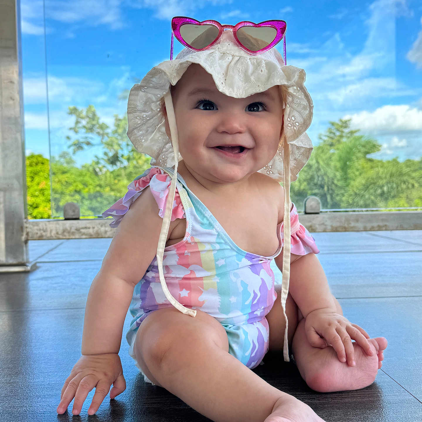Zaenhyra is registered to the contest to win money with this photo: baby, barefoot, blue_sky, child, cute, daylight, floor, greenery, happy, hat, heart_shaped, infant, outdoor, portrait, sitting, smiling, sun_hat, sunglasses, swimsuit, unicorn_pattern