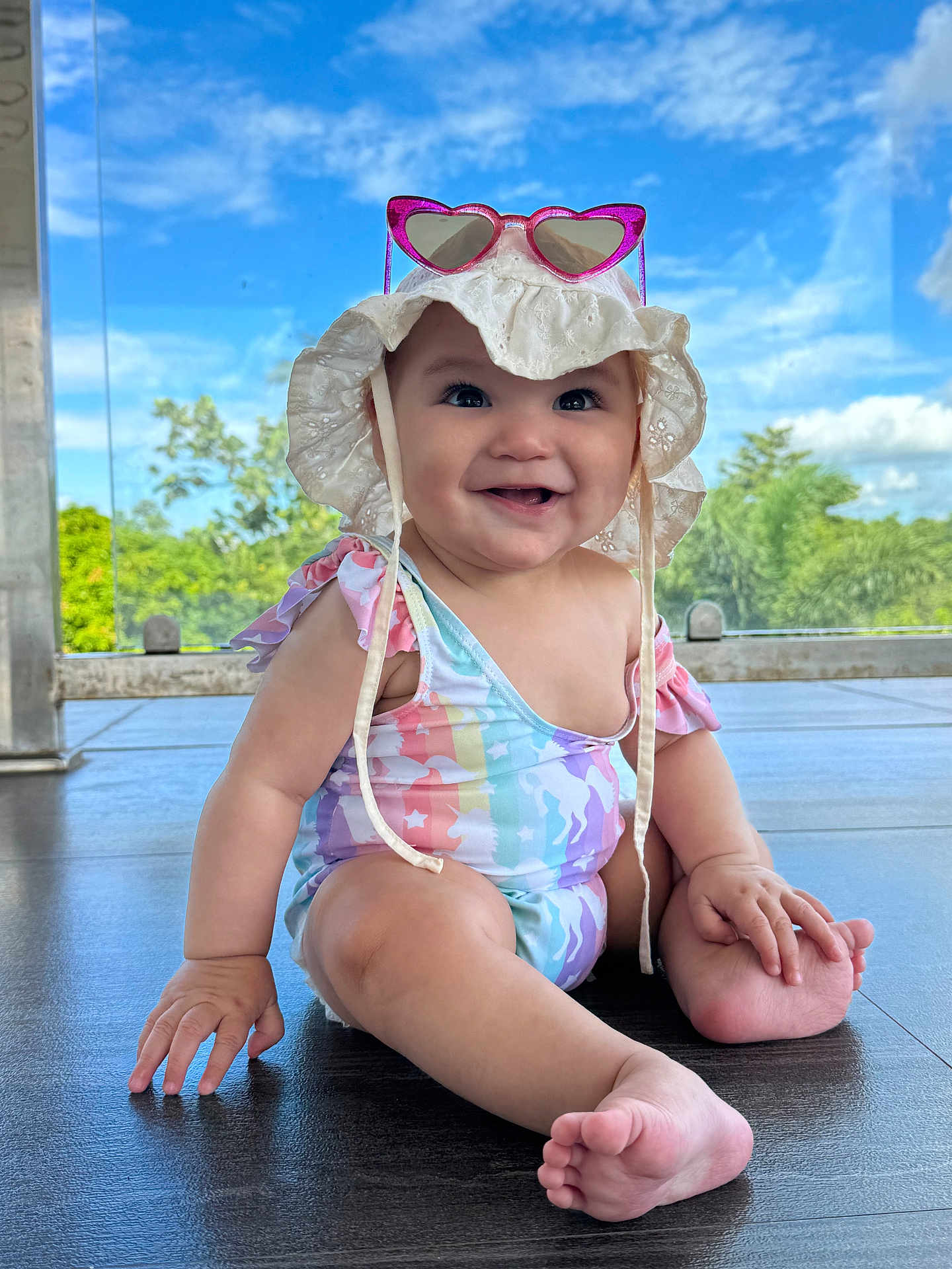 Zaenhyra is registered to the contest to win money with this photo: baby, child, smiling, sitting, floor, hat, sunglasses, heart_shaped, swimsuit, unicorn_pattern, outdoor, daylight, blue_sky, greenery, happy, cute, infant, portrait, sun_hat, barefoot