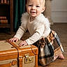 baby, child, toddler, smiling, portrait, indoors, suitcase, luggage, wooden_floor, bookshelf, books, curtains, statue, home_decor, fashion, sweater, skirt, crawling, vintage, travel