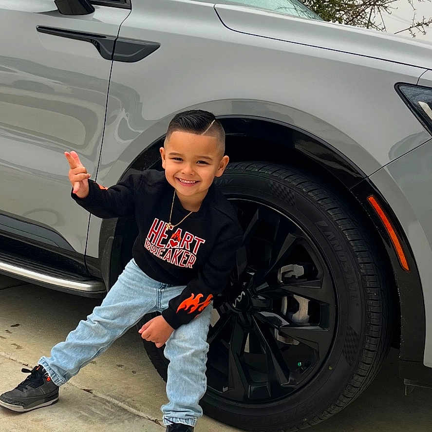 Ismael Gomez joined the competition — help win amazing prizes! black_sweatshirt, boy, car, casual_clothing, child, concrete, driveway, flames, light_blue_jeans, outdoor, overcast, peace_sign, portrait, smiling, sneakers, suv, tire, tree, vehicle, wheel
