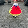 toddler, child, costume, snow_white, red_bow, yellow_tutu, walking, asphalt, stroller, brick_wall, car, tire, outdoor, back_view, curly_hair, cape, sidewalk, vehicle, parking_lot, playful