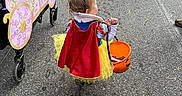Drue is registered to the contest to win money with this photo: toddler, child, costume, snow_white, yellow_tutu, red_bow, pumpkin_bucket, candy, stroller, carriage_decoration, street, pavement, people, legs, shoes, outdoor, festival, halloween, walking, back_view