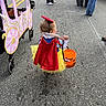 toddler, child, costume, snow_white, yellow_tutu, red_bow, pumpkin_bucket, candy, stroller, carriage_decoration, street, pavement, people, legs, shoes, outdoor, festival, halloween, walking, back_view