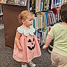 Drue is registered to the contest to win money with this photo: toddler, child, library, bookshelf, books, dress, pumpkin, costume, indoor, carpet, footwear, socks, person, motion_blur, short_hair, curly_hair, earrings, young_child, reading, aisle