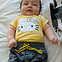 baby, infant, child, yellow_shirt, hello_kitty, grey_pants, batman_logo, drawstring, bed, blanket, lying_down, face, arms, cute, soft_lighting, indoor, person, clothing, comfort, smile