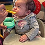 baby, feeding, high_chair, spoon, food, container, gray_clothing, elephant_print, indoor, curious, child, hand, person, table, cushion, plaid, red_letters, homedecor, cute, infant