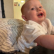 Rayden is registered to the contest to win money with this photo: baby, infant, smiling, teeth, highchair, feeding_tray, white_shirt, patterned_pillow, ear, eye, face, indoor, party_banner, wall, cushion, close_up, portrait, happy, drooling, cheek