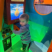 Mylin is registered to the contest to win money with this photo: amusement, arcade, child, clothing, colorful, curious, entertainment, game, green_light, indoor, neon_light, pants, play, reflection, screen, sneakers, standing, submarine, toddler, toy