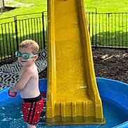 Mylin is registered to the contest to win money with this photo: child, toddler, swimming_pool, slide, yellow_slide, goggles, water, red_shorts, outdoor, playground, summer, fun, play, green_goggles, bare_chest, grass, fence, toy, smile, person