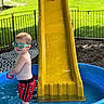 bare_chest, child, fence, fun, goggles, grass, green_goggles, outdoor, person, play, playground, red_shorts, slide, smile, summer, swimming_pool, toddler, toy, water, yellow_slide