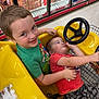 Mylin is registered to the contest to win money with this photo: child, children, shopping_cart, toy_car, yellow, smiling, store, aisle, supermarket, steering_wheel, green_shirt, red_shirt, shorts, playful, happy, indoor, person, retail, fun, face