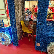 Maëlan a rejoint le concours — aidez-le/la à gagner de superbes lots ! blue_tinsel, child, curious, decorations, festive, holiday_decor, indoor, mall, people, public_space, red_carpet, seated, shopping, shopping_cart, silver_tinsel, tinsel, toddler, toy_train, woman, yellow_chair