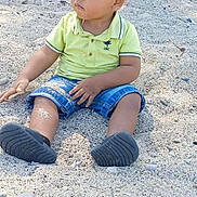 Maëlan participe au concours pour gagner de l'argent avec cette photo : child, toddler, sand, outdoor, sitting, shorts, polo_shirt, green_shirt, black_shoes, legs, face, nature, trees, fence, daylight, casual_clothing, young_child, ground, curious, playful
