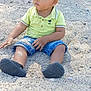 child, toddler, sand, outdoor, sitting, shorts, polo_shirt, green_shirt, black_shoes, legs, face, nature, trees, fence, daylight, casual_clothing, young_child, ground, curious, playful