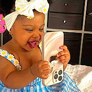 Arabelle Olivia is registered to the contest to win money with this photo: toddler, child, smiling, happy, phone, smartphone, dress, blue, white, bow, daisies, hair_accessory, bed, indoor, furniture, cabinet, cute, playful, holding, portrait