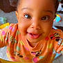 toddler, child, smiling, face, eyes, hair_ties, orange_shirt, cartoon_characters, polka_dot, tablecloth, happy, indoor, closeup, cute, person, playful, bright_colors, curly_hair, baby, expression