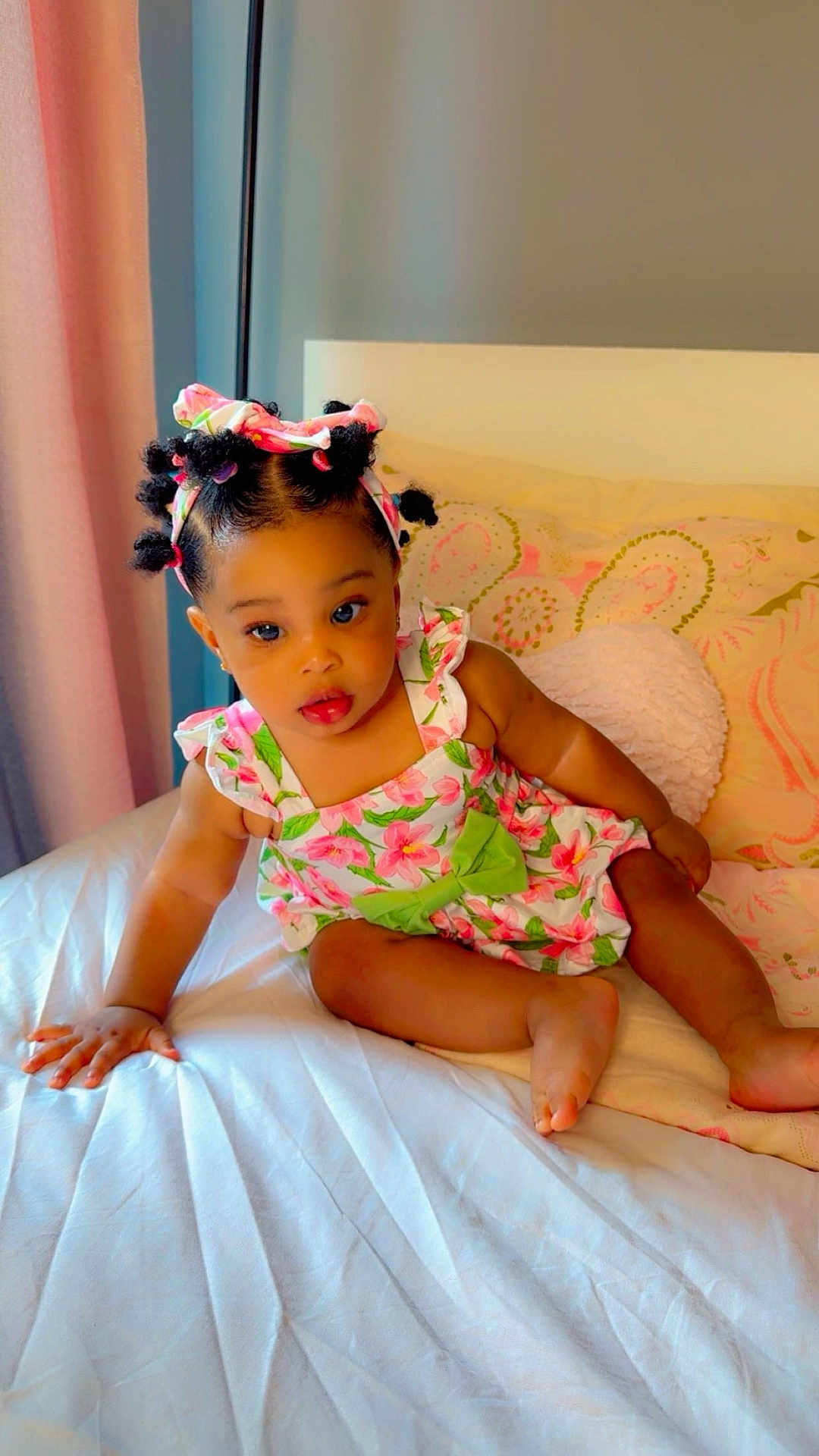 Arabelle Olivia joined the competition — help win amazing prizes! baby, toddler, child, face, hair, headband, floral_pattern, dress, green_bow, bed, pillow, pink, white_sheet, indoors, curious, cute, sitting, skin, hand, foot