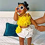 toddler, child, sunglasses, yellow_top, white_skirt, purse, bed, quilt, hand, adult, indoor, fashion, footwear, sandals, smiling, cute, hair_buns, portrait, home, decor