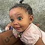 baby, carpet, child, closeup, curly_hair, cute, earring, hand, holding, indoor, infant, person, pink_clothing, portrait, skin, smile, soft_toy, tattoo, wide_eyes, young_child