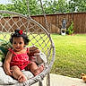 baby, child, cushion, cute, fence, flower_hair, footwear, garden, grass, greenery, hand, hanging_chair, infant, outdoor, person, porch, red_dress, relaxed, smiling, sunny