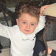 Gabriel is registered to the contest to win money with this photo: toddler, child, boy, curly_hair, blue_eyes, white_shirt, black_pants, sitting, floor, tile_floor, chair, hand, indoor, casual_clothing, person, cute, portrait, looking_at_camera, young_child, home