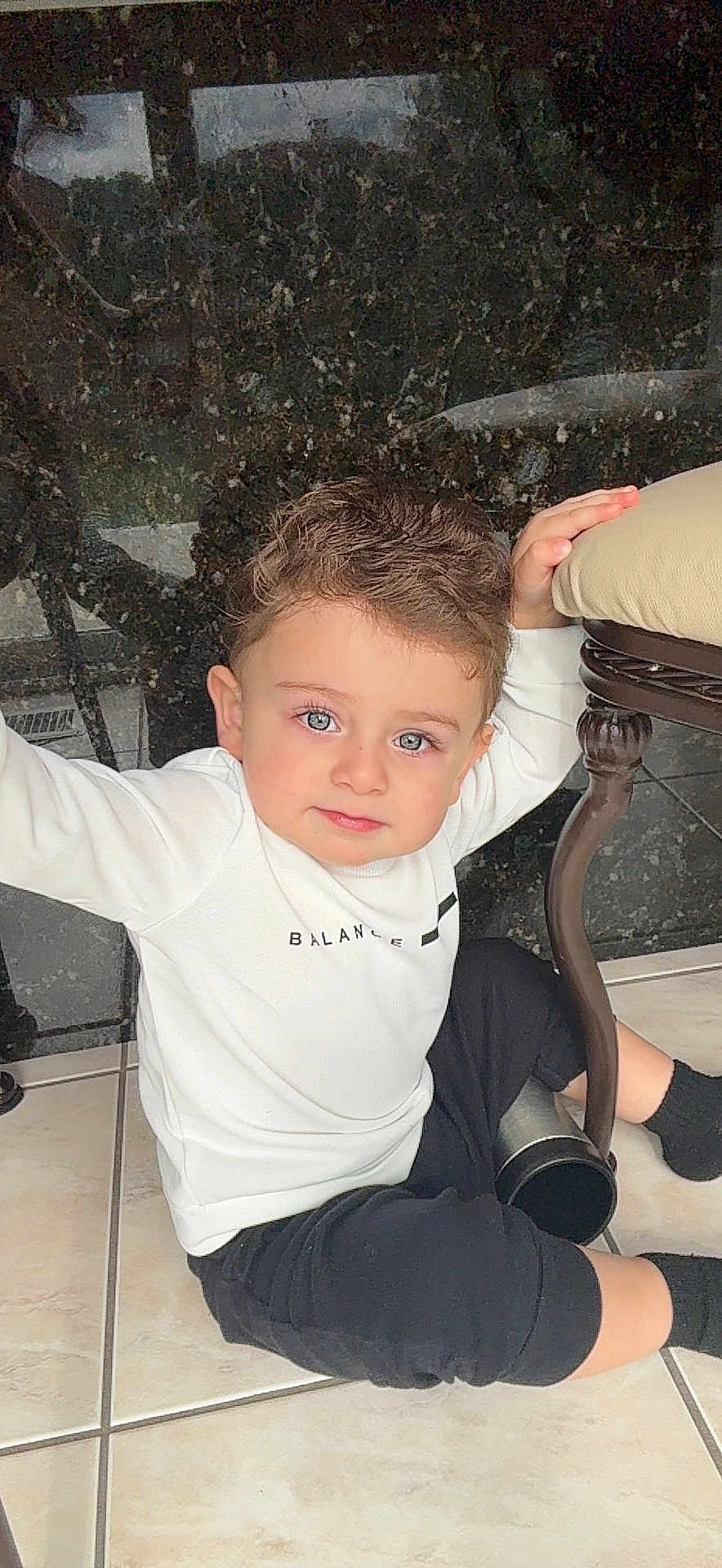 Gabriel is registered to the contest to win money with this photo: toddler, child, boy, curly_hair, blue_eyes, white_shirt, black_pants, sitting, floor, tile_floor, chair, hand, indoor, casual_clothing, person, cute, portrait, looking_at_camera, young_child, home