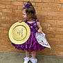 child, girl, purple_dress, tiara, award, certificate, brick_wall, side_view, white_shoes, socks, curly_hair, concrete_floor, doll, standing, portrait, celebration, costume, young_child, outdoor, happy