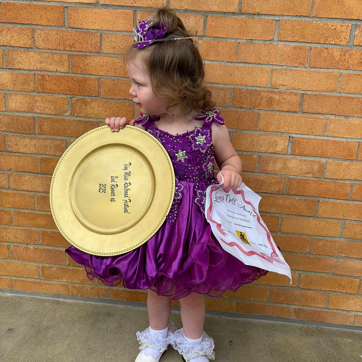 Azah is registered to the contest to win money with this photo: award, brick_wall, celebration, certificate, child, concrete_floor, costume, curly_hair, doll, girl, happy, outdoor, portrait, purple_dress, side_view, socks, standing, tiara, white_shoes, young_child