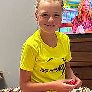 Izabell is registered to the contest to win money with this photo: bedroom, bedspread, casual_clothing, child, colorful, dresser, furniture, hair_bun, happy, holding_object, indoor, person, portrait, relaxed, sitting, smile, television, woman, yellow_tshirt, young