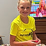 bedroom, bedspread, casual_clothing, child, colorful, dresser, furniture, hair_bun, happy, holding_object, indoor, person, portrait, relaxed, sitting, smile, television, woman, yellow_tshirt, young