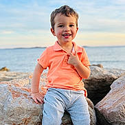 Ayden a rejoint le concours — aidez-le/la à gagner de superbes lots ! child, toddler, boy, smiling, orange_polo, grey_pants, rock, rocks, ocean, water, sky, sunset, outdoor, nature, happy, portrait, sitting, casual_clothing, daylight, coast