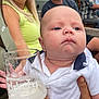 baby, infant, person, adult, holding, glass, beer, drink, white_shirt, polo_shirt, hand, face, expression, outdoor, social_gathering, people, seated, casual_clothing, beverage, portrait