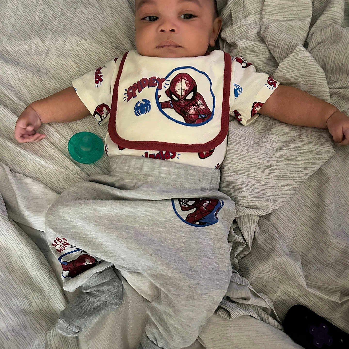 Treyson is registered to the contest to win money with this photo: baby, bed, blanket, casual, child, clothing, cute, fabric, indoor, infant, lying_down, pacifier, person, portrait, relaxed, remote_control, sleepwear, socks, spiderman, young