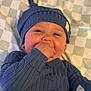 baby, infant, blue_clothing, hat, hand_in_mouth, blanket, checkered_pattern, cute, child, smiling, lying_down, face, skin, one_person, indoors, soft_lighting, close_up, portrait, happy, young_child