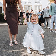 Judith a rejoint le concours — aidez-le/la à gagner de superbes lots ! toddler, child, adult_hand, white_dress, pacifier, outdoor, group_of_people, building, pavement, shoes, woman, holding_hand, crowd, dress, summer, daylight, people, event, casual_clothing, urban