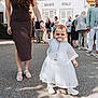 toddler, child, adult_hand, white_dress, pacifier, outdoor, group_of_people, building, pavement, shoes, woman, holding_hand, crowd, dress, summer, daylight, people, event, casual_clothing, urban