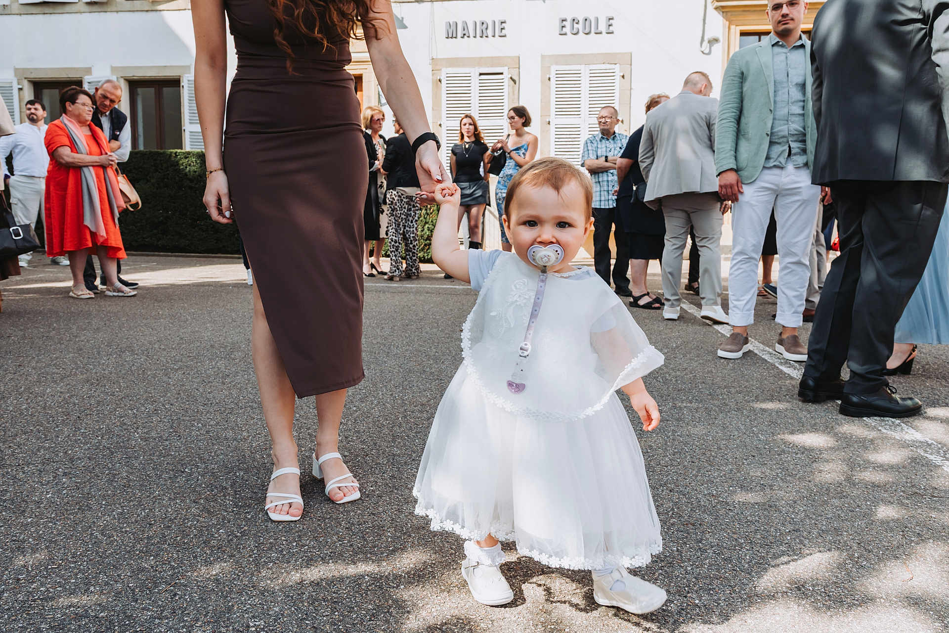 Judith a rejoint le concours — aidez-le/la à gagner de superbes lots ! toddler, child, adult_hand, white_dress, pacifier, outdoor, group_of_people, building, pavement, shoes, woman, holding_hand, crowd, dress, summer, daylight, people, event, casual_clothing, urban