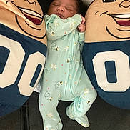 Jaziya is registered to the contest to win money with this photo: baby, sleeping, onesie, plush_pillow, cartoon_face, smile, bedroom, child, infant, cute, cozy, blue, green, patterned_clothing, soft_toys, comfort, relaxation, rest, indoor, person