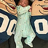 baby, sleeping, onesie, plush_pillow, cartoon_face, smile, bedroom, child, infant, cute, cozy, blue, green, patterned_clothing, soft_toys, comfort, relaxation, rest, indoor, person