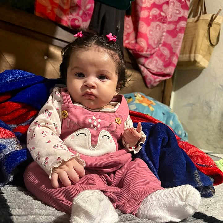 Layla is registered to the contest to win money with this photo: baby, child, infant, sitting, bed, blanket, pink_outfit, bunny_outfit, hair_bows, smiling, portrait, indoors, bedroom, colorful, clothing, cushion, toy, tote_bag, natural_light, cute