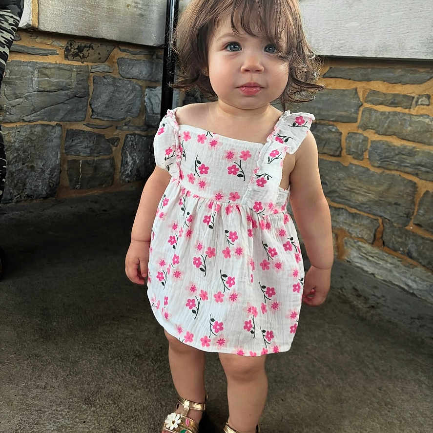 Alaia is registered to the contest to win money with this photo: casual_clothing, child, concrete_floor, curly_hair, cute, expression, fashion, floral_dress, footwear, girl, innocent, outdoor, person, portrait, sandals, standing, stone_wall, summer, toddler, young