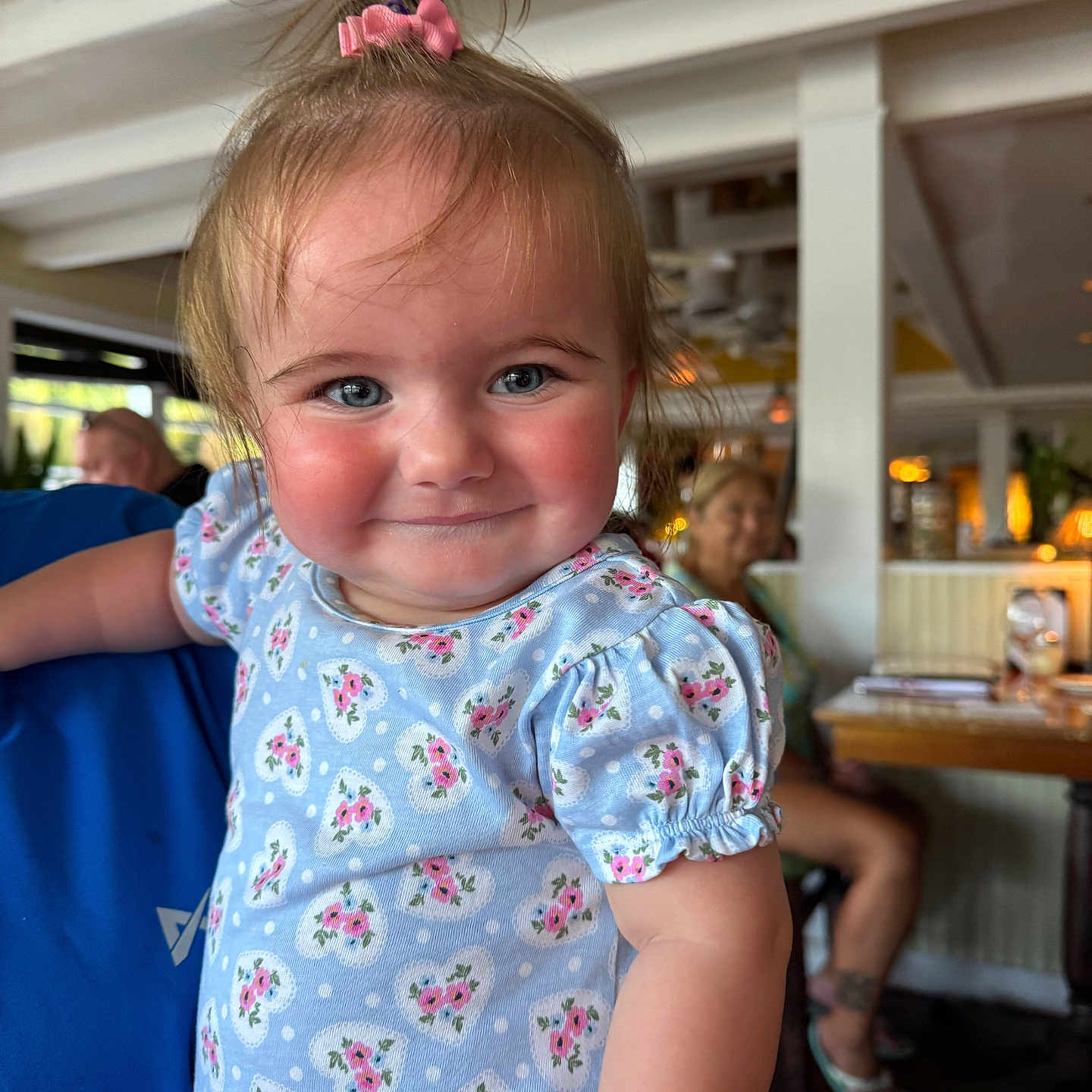 Marlow is registered to the contest to win money with this photo: background, blue_dress, casual, child, cute, face, floral_pattern, funny_hair, hair_tie, happy, holding, indoor, people, person, portrait, restaurant, rosy_cheeks, short_sleeves, smile, toddler