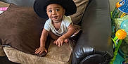 Malano joined the competition — help win amazing prizes! baby, child, hat, cowboy_hat, couch, pillow, brown_pillow, black_couch, wooden_floor, indoor, smiling, happy, cute, seated, toys, floor, person, infant, playful, cozy