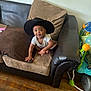 baby, child, hat, cowboy_hat, couch, pillow, brown_pillow, black_couch, wooden_floor, indoor, smiling, happy, cute, seated, toys, floor, person, infant, playful, cozy