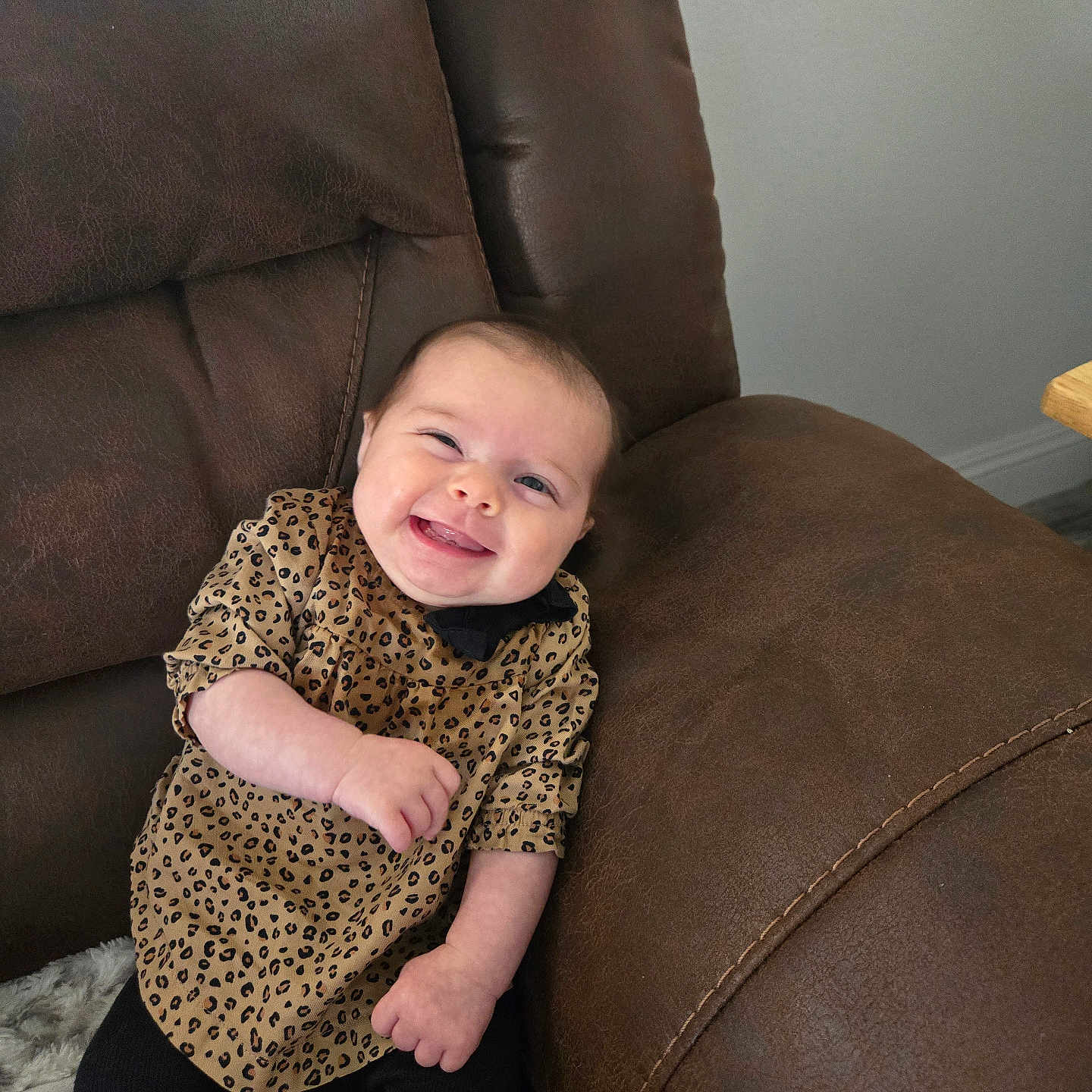 Maeve is registered to the contest to win money with this photo: baby, black_pants, brown_leather_couch, child, clothing, comfort, cute, furniture, happy, home, indoor, infant, leopard_print, person, portrait, relaxed, seat, sitting, smiling, young