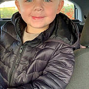 Lucas a rejoint le concours — aidez-le/la à gagner de superbes lots ! child, boy, smile, blue_eyes, puffer_jacket, black_jacket, car_interior, seat, window, outdoor_background, nature, greenery, orange_flowers, portrait, happy, casual_clothing, young, person, face, head