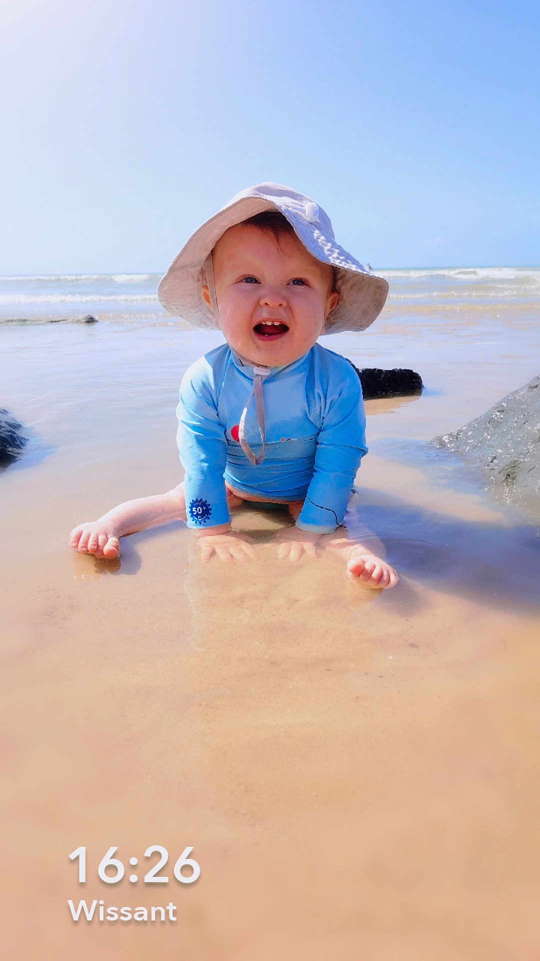 Lyzio participe au concours pour gagner de l'argent avec cette photo : baby, child, beach, water, sun_hat, blue_sky, ocean, waves, sun_protection, happy, smiling, sitting, shallow_water, summer, outdoor, sand, sunny, playful, cute, vacation