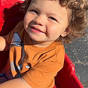 Brantley is registered to the contest to win money with this photo: child, toddler, curly_hair, smile, orange_tshirt, grey_pants, red_wagon, outdoor, sunlight, happy, play, daylight, seatbelt, casual_clothing, cute, person, fun, park, young_child, playtime