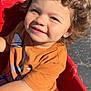 child, toddler, curly_hair, smile, orange_tshirt, grey_pants, red_wagon, outdoor, sunlight, happy, play, daylight, seatbelt, casual_clothing, cute, person, fun, park, young_child, playtime