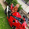 child, toddler, motorcycle, red_motorcycle, grass, lawn, house, porch, cushions, greenery, outdoor, sidewalk, curly_hair, orange_clothing, pumpkin_pattern, person, vehicle, toy, daytime, casual
